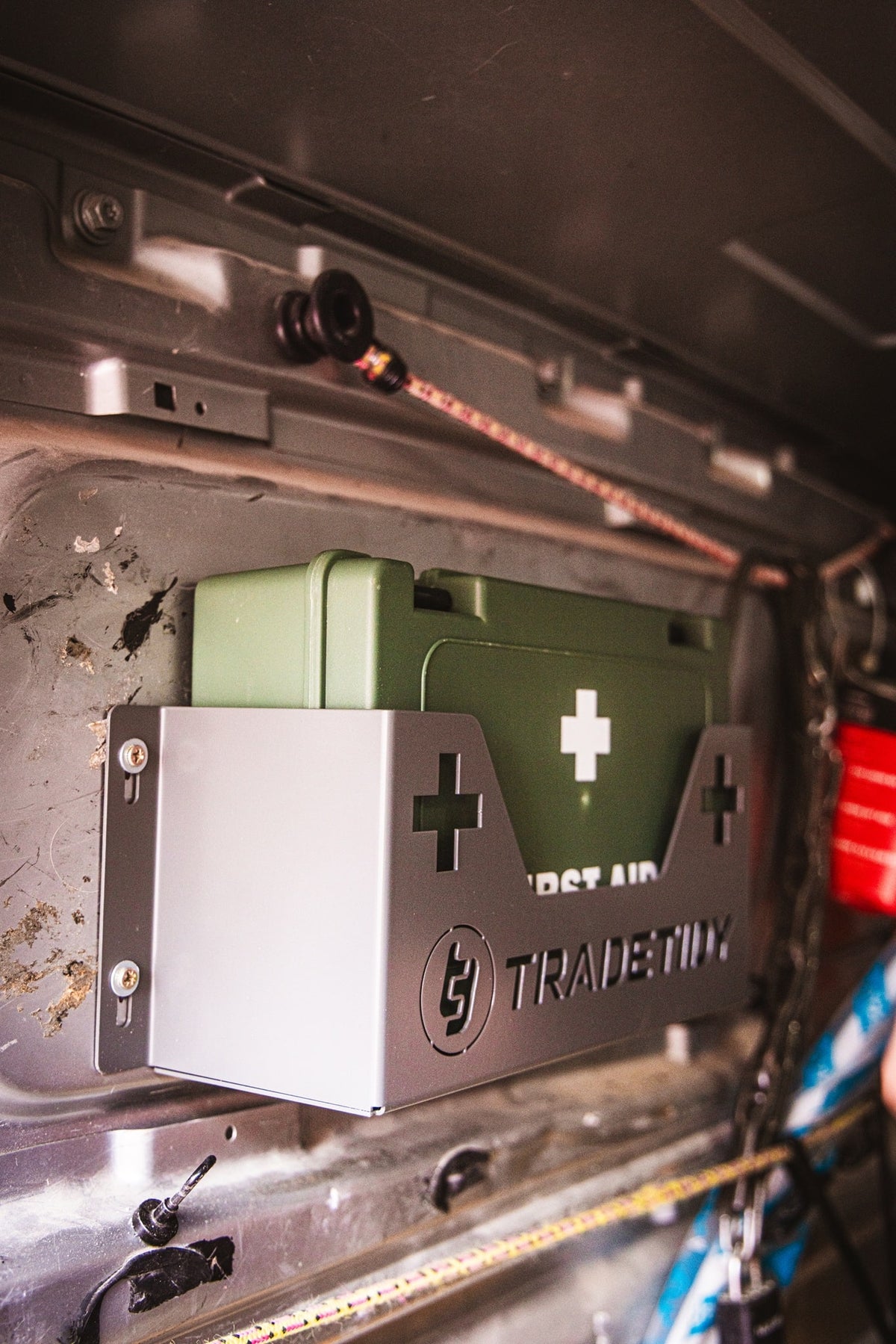 Green first aid kit in a grey first aid kit holder with 'TradeTidy' branding on a workshop van's wall