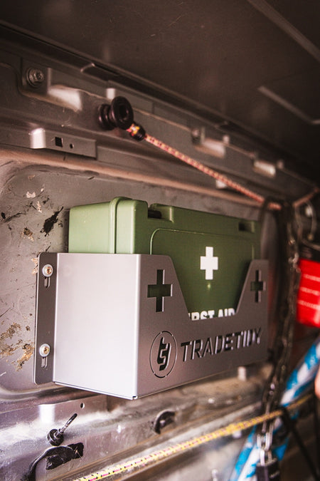 Green first aid kit in a grey first aid kit holder with 'TradeTidy' branding on a workshop van's wall