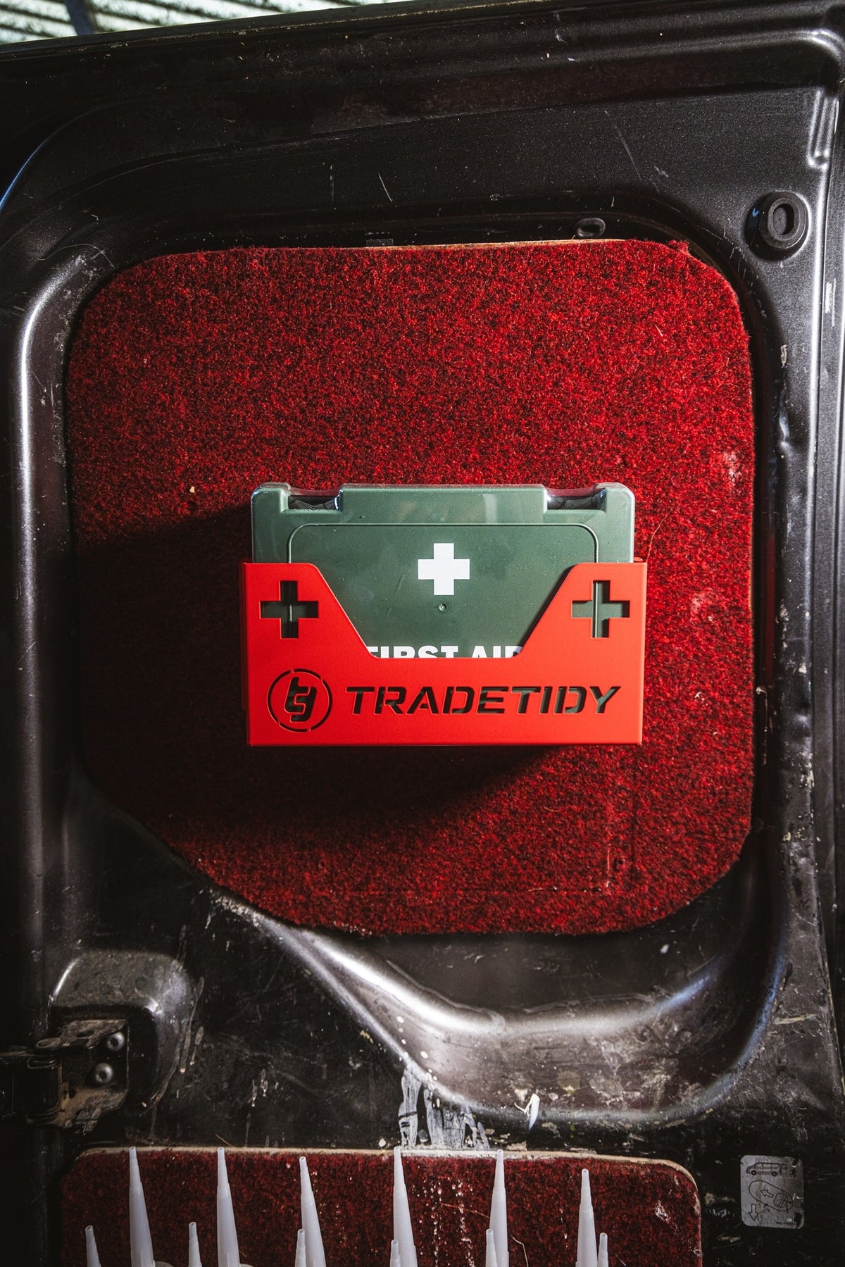 Green First aid kit in a red first aid kit holder on a red textured surface with 'TradeTidy' branding.