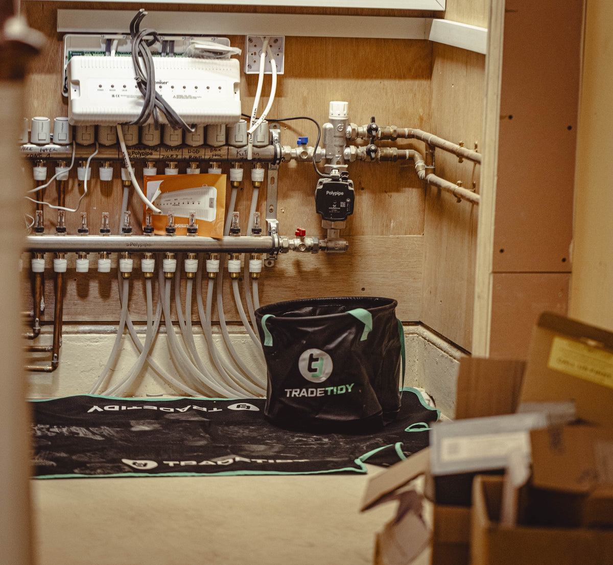Mechanical room with pipes and valves, TradeTidy bag on a mat, and boxes in the foreground.