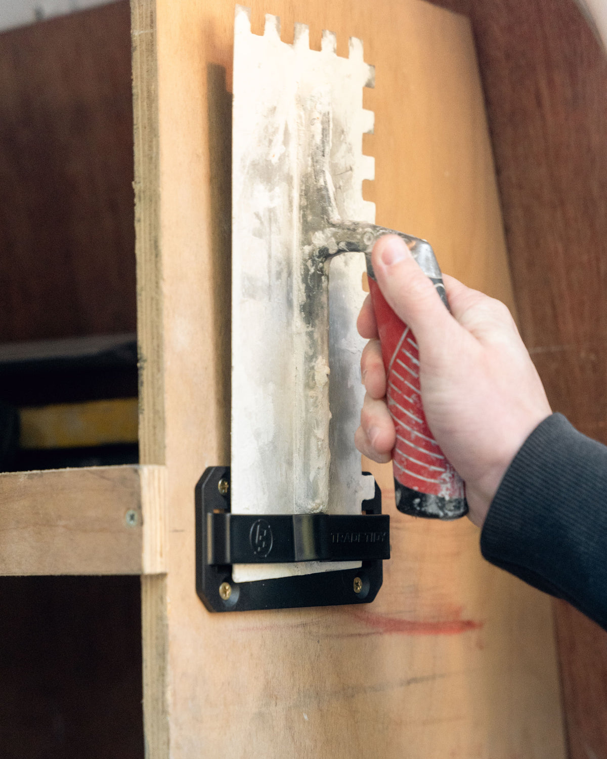 TradeTidy - hand holding a trowel in a black trowel holder hanging on a wooden wall